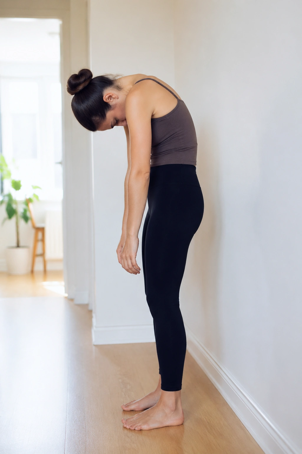 Person performing standing roll-down near a wall, knees bent, spine curling forward